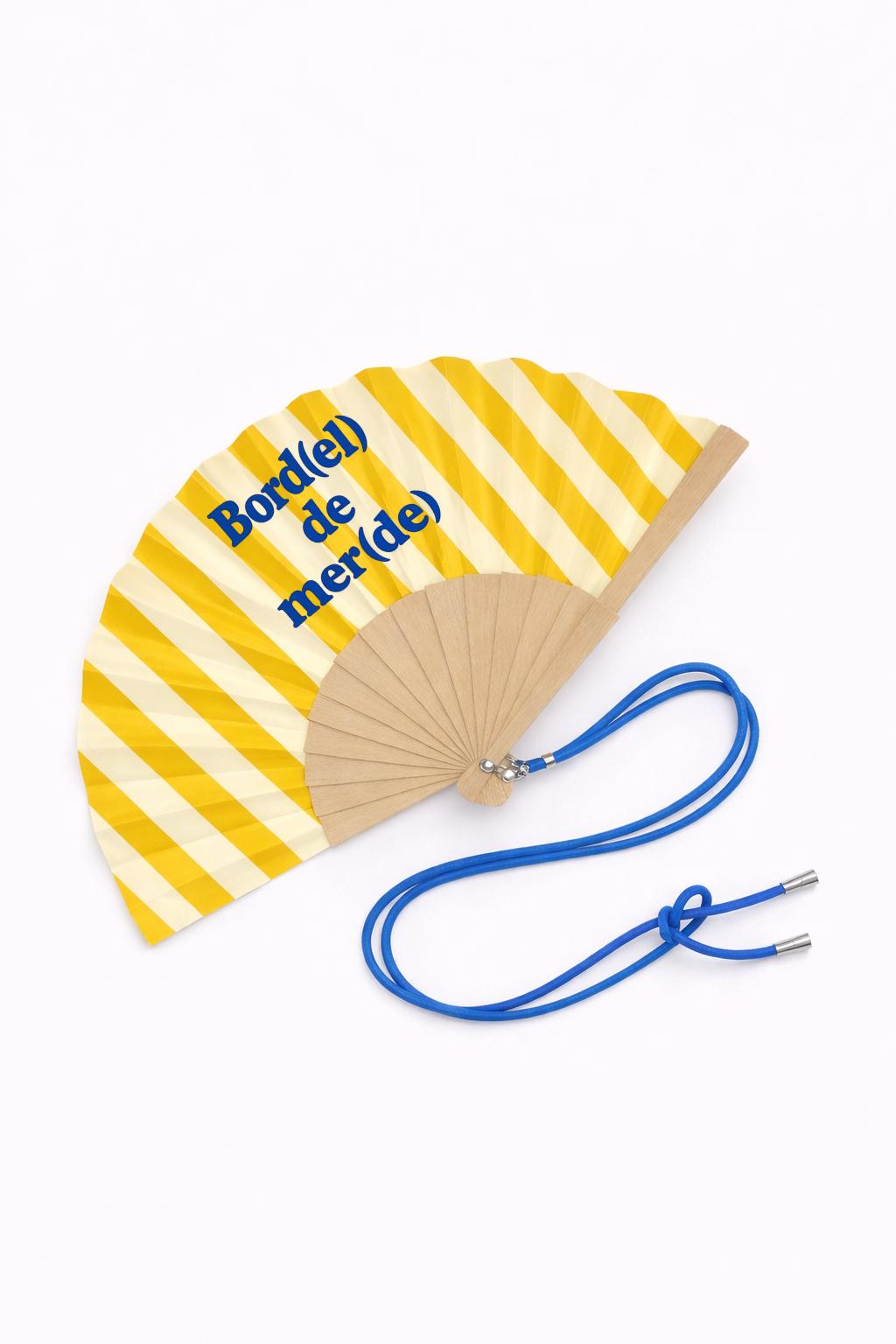 Éventail pliable en bois et tissu à rayures jaunes avec le message « Bord(el) de mer(de) ».
Doté d’une bandoulière bleue ajustable pour l’avoir toujours à portée de main pendant l’été.
